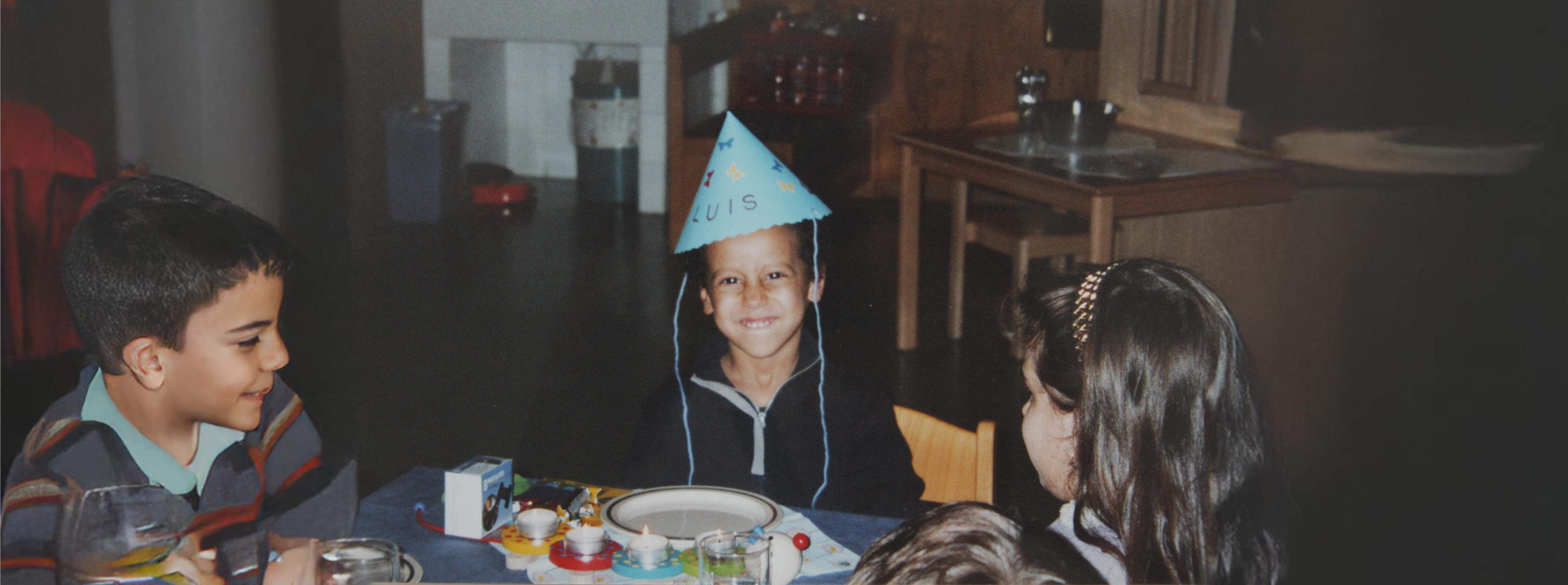 Luis as a child, wearing a handmade party hat at a birthday table.